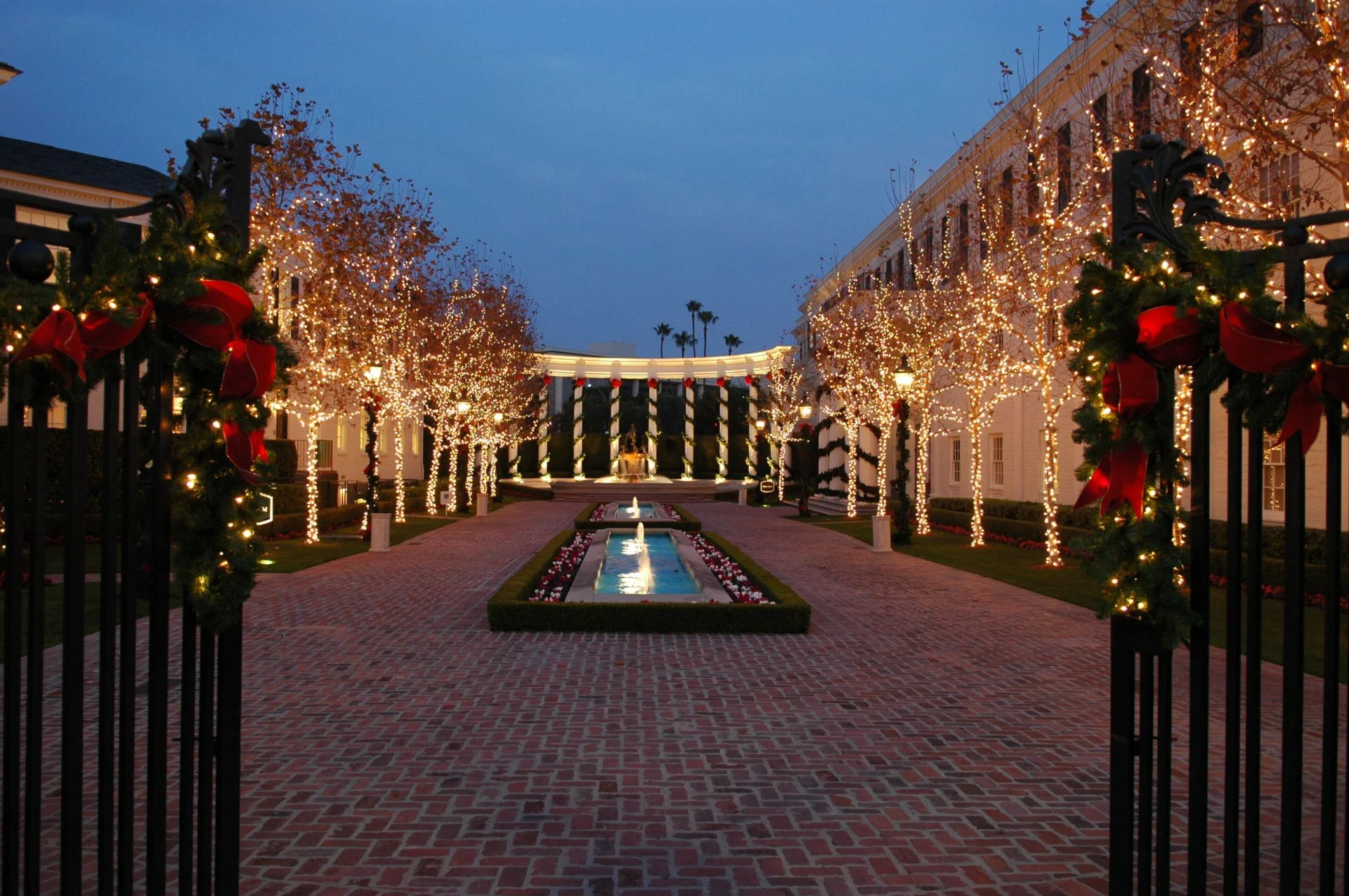 Tree-lined courtyard walkway with fountains, lit at dusk